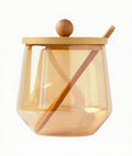 A translucent amber glass jar with a light natural wood lid featuring a round wooden knob and a wooden spoon resting inside, shown on a solid white background.
